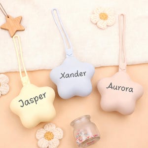 May include: Three star-shaped pacifier holders in pastel colors: yellow, blue, and pink. Each holder has a name printed on it: Jasper, Xander, and Aurora. A small glass jar with colorful sprinkles and crocheted flowers are also in the image.