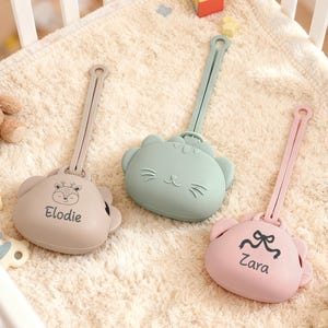 May include: Three baby pacifier holders in beige, mint green, and pink, each with a different animal face design. The beige holder has the name "Elodie" printed on it, and the pink holder has the name "Zara". The holders are on a soft, cream-colored blanket.