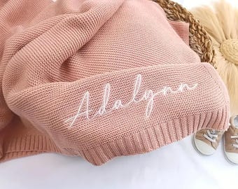 Puede incluir: Una manta de bebé de punto rosa polvoriento con el nombre "Adalynn" bordado en escritura blanca. La manta está doblada en una cesta tejida, con un par de zapatos pequeños de color canela visibles.