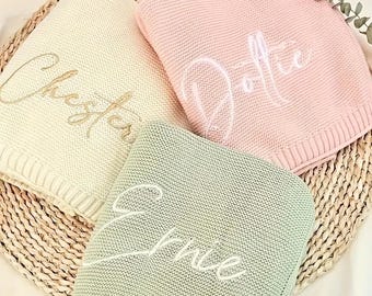Puede incluir: Tres mantas de bebé plegadas en crema, rosa y verde salvia, cada una bordada con un nombre. Las mantas se muestran sobre una estera tejida. La manta crema tiene 'Chester' bordado en dorado.