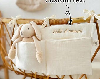 May include: A cream-colored baby crib organizer with two pockets and a stuffed bunny. The organizer has the text "Bébé d'amour" embroidered on it. The crib is made of woven wood.