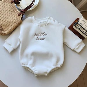 Op de afbeelding: Een witte baby-onesie met lange mouwen en een ronde halslijn. De woorden "little love" zijn in een cursief lettertype op de voorkant geborduurd. De onesie heeft drukknopen op de schouder en een gerimpelde onderkant.