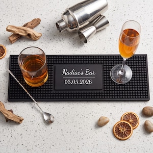 May include: A rectangular black rubber bar mat with a textured nub surface, featuring a central plaque printed with the text "Nadiac's Bar" and "03.05.2026". The mat is accompanied by a cocktail shaker, a jigger, a mixing spoon, and two glasses.