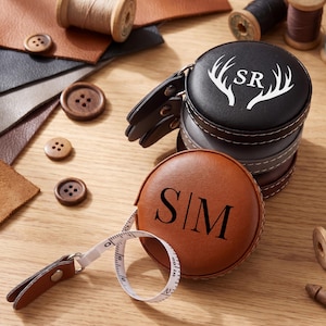 Op de afbeelding: Een stapel gepersonaliseerde leren meetlinten in bruin, grijs en zwart. Het bruine meetlint is open en toont de meetlint. Het zwarte meetlint heeft de initialen "SR" en geweien. Houten knopen en leerresten op de achtergrond.