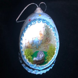 May include: A blue and white egg-shaped ornament with a Peter Rabbit illustration inside. The ornament is decorated with blue and black lace trim and has a silver hanger.