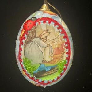 May include: An oval-shaped hanging ornament featuring a central illustration of two rabbits in a garden setting. The image is framed by a red braided trim and accented with a small red ladybug. A gold-colored hanging loop is attached at the top.