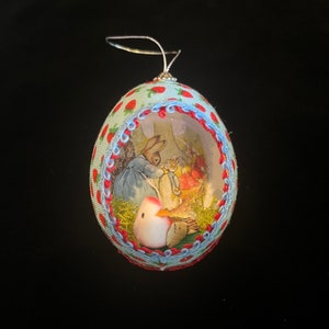 May include: A blue and white egg-shaped ornament with a red and white ribbon around it. The ornament has a clear plastic window that shows a picture of a rabbit and a bird inside.