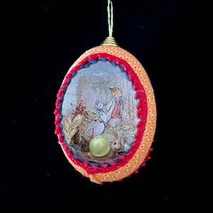 May include: A handmade Christmas ornament shaped like an egg with a red and orange fabric exterior. The ornament features a small, detailed scene inside with a snail, a bird, and a figure in a forest setting.