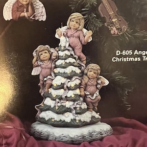 May include: A ceramic Christmas tree figurine with four angels, a violin ornament, and a base that holds a music box. The tree is decorated with white snow and gold accents. The base is labeled "D-604 Christmas Tree Base".