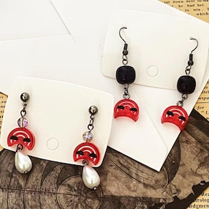 May include: A pair of handmade earrings featuring a red, semi-circular charm with a black design, a black square bead, and a white teardrop-shaped pearl. The earrings are on white earring display cards.