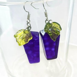 May include: A pair of dangle earrings featuring a grape cluster design. Each earring has a purple, rectangular grape cluster with a green leaf accent. The earrings are suspended from silver-colored fish hook ear wires.