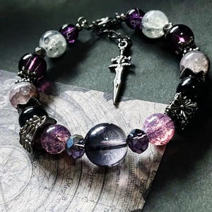 May include: A beaded bracelet featuring a variety of purple, clear, and black beads. The bracelet includes a silver-colored sword charm and decorative metal accents. The beads vary in size and texture, creating a unique and detailed accessory.