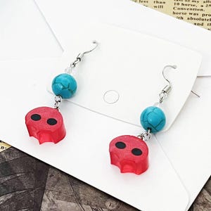 Op de afbeelding: Hangende oorbellen met rode schedelbedels met zwarte oogdetails, gecombineerd met turkooizen kralen. De oorbellen, met zilverkleurige haken, worden gepresenteerd op een witte kaart.
