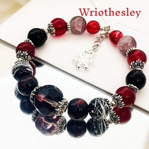 Puede incluir: Un brazalete de cuentas redondas en tonos rojos, negros y grises. El brazalete incluye un dije de copo de nieve y la palabra "Wriothesley" en texto rojo. Las cuentas tienen detalles plateados.