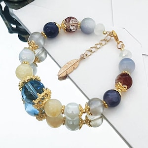 May include: A beaded bracelet featuring a variety of colorful beads in shades of blue, yellow, and gray. The bracelet includes gold-colored accents and a feather charm. The beads vary in size and texture, creating a unique and eye-catching accessory.