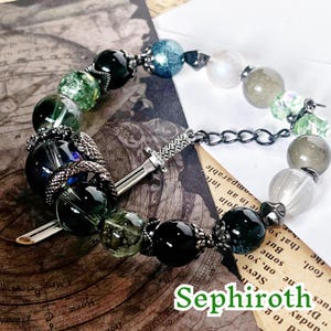 Puede incluir: Una pulsera de cuentas con cuentas de piedras preciosas verdes, negras y transparentes, y un dije de espada plateada. La pulsera está conectada por una cadena y tiene la palabra "Sephiroth".