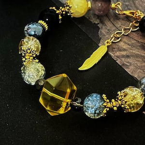 Puede incluir: Una pulsera de cuentas con una variedad de piedras preciosas coloridas y detalles dorados. La pulsera incluye cuentas amarillas, negras y azules, con un dije de pluma dorada. El diseño incorpora cuentas facetadas y redondas.