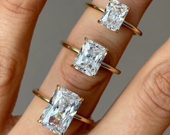 May include: Three gold rings with rectangular, clear gemstones are displayed on a finger. The rings have a simple, elegant design with a thin gold band. The gemstones are cut in a rectangular shape, and they sparkle in the light.