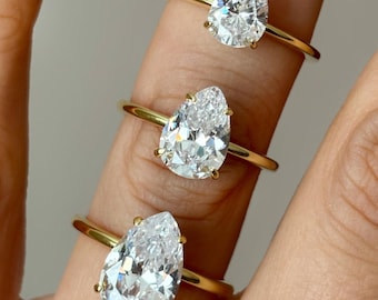May include: Three gold rings, each featuring a pear-shaped diamond. The diamonds are clear and sparkling, set on delicate gold bands. The rings are displayed on a person's finger, showcasing the jewelry's design and brilliance.