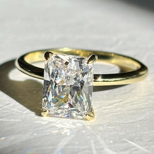 Può includere: Un anello di fidanzamento in oro con un diamante taglio rettangolare. Il diamante è tenuto in posizione da quattro griffe e la fascia è liscia e lucida. L'anello è appoggiato su una superficie bianca, con la luce del sole che crea un'ombra.