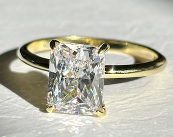 May include: A radiant-cut diamond engagement ring with a gold band. The large, rectangular diamond is set with four prongs, and the gold band is smooth and polished. The ring is displayed on a white surface, with sunlight casting a shadow.