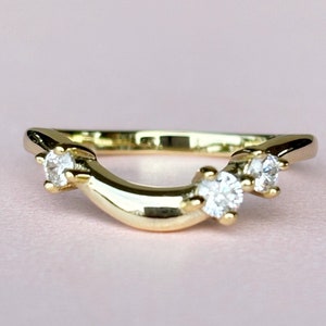 Può includere: Un anello curvo dorato con tre pietre preziose rotonde e trasparenti. L'anello ha una finitura liscia e lucida ed è posto su uno sfondo rosa tenue. Le pietre preziose sono equidistanti lungo la curva dell'anello.