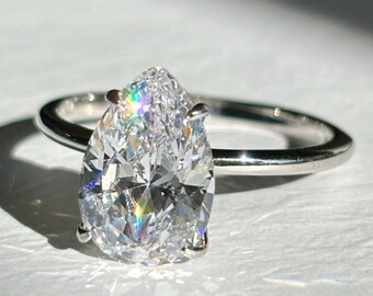 May include: A pear-shaped diamond engagement ring with a silver band. The large, clear diamond sparkles with rainbow reflections. The ring is set against a white background, with shadows adding depth.