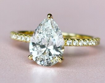 May include: A gold ring with a pear-shaped diamond center stone. The band is also adorned with small diamonds. The ring is set against a soft pink background, highlighting the sparkle of the gemstones.