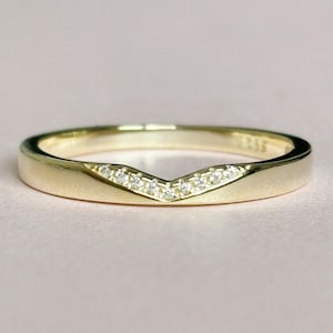 Op de afbeelding: Een goudkleurige ring met een V-vormig ontwerp, bezet met kleine, heldere edelstenen. De ring heeft een gladde, gepolijste band en een delicate, elegante uitstraling. De ring is waarschijnlijk gemaakt van goud en is bedoeld om aan een vinger te worden gedragen.