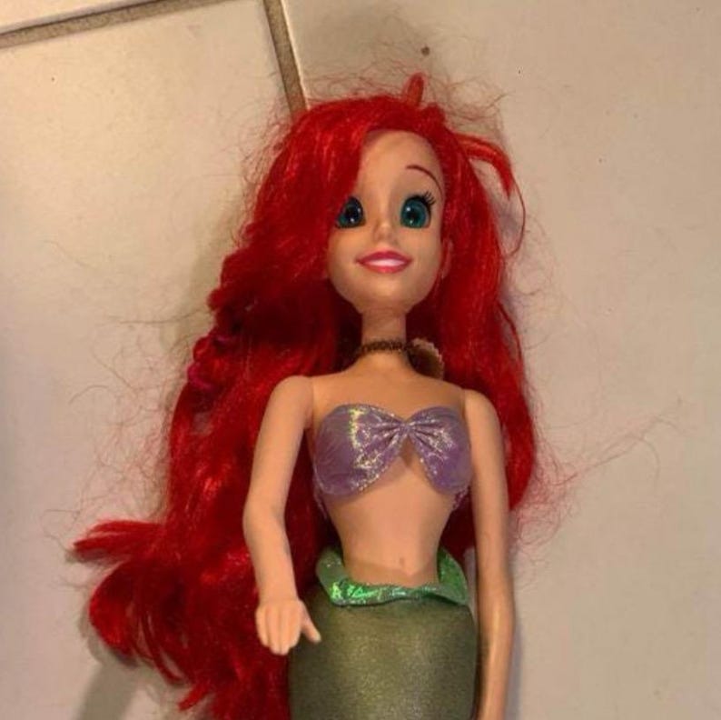 Pode incluir: Uma boneca sereia com cabelo comprido e vermelho vibrante. A boneca tem um top roxo e uma cauda verde. A boneca tem uma express&atilde;o sorridente e usa um colar preto. A boneca &eacute; uma personagem popular de um filme da Disney.