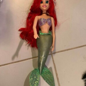 Pode incluir: Boneca de sereia com longos cabelos ruivos, top roxo em forma de concha e cauda verde brilhante. A boneca tem uma express&atilde;o sorridente e &eacute; um brinquedo popular para crian&ccedil;as.
