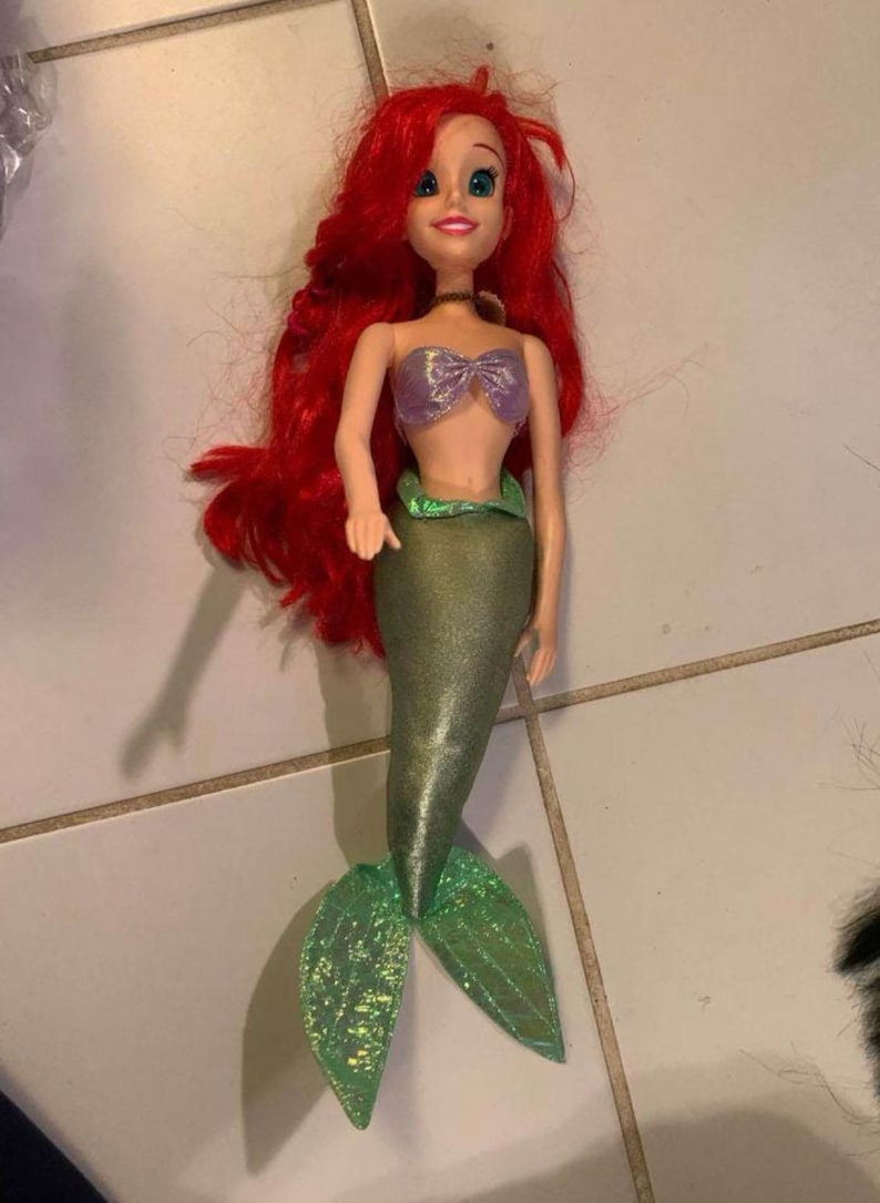 Pode incluir: Boneca de sereia com longos cabelos ruivos, top roxo em forma de concha e cauda verde brilhante. A boneca tem uma express&atilde;o sorridente e &eacute; um brinquedo popular para crian&ccedil;as.