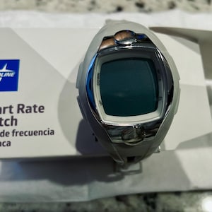 Pu&ograve; includere: Un orologio Medline per la frequenza cardiaca grigio e argento con display digitale. L'orologio ha pulsanti con le etichette "Up", "Down" e "Mode". L'orologio &egrave; sopra una scatola bianca con il logo Medline e le parole "Heart Rate Watch" e "Reloj de frecuencia cardiaca".