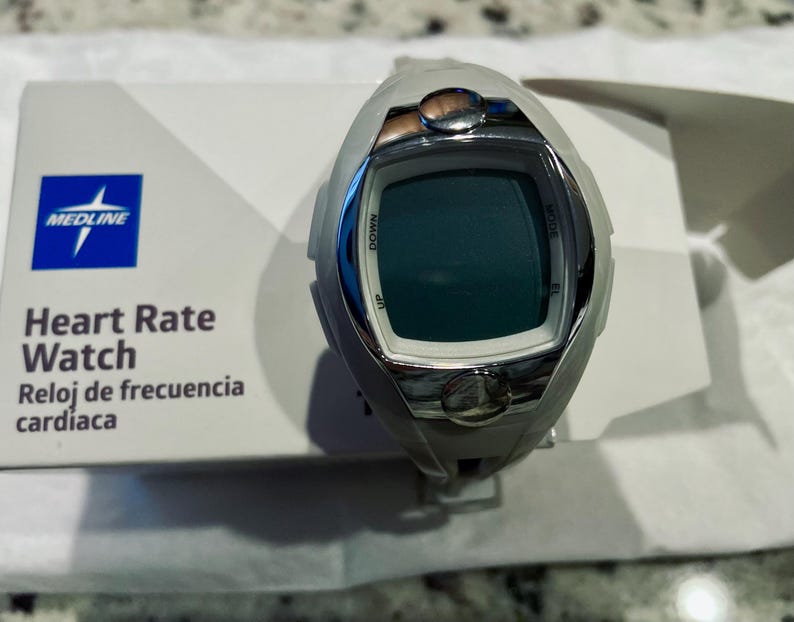 Pu&ograve; includere: Un orologio Medline per la frequenza cardiaca grigio e argento con display digitale. L'orologio ha pulsanti con le etichette "Up", "Down" e "Mode". L'orologio &egrave; sopra una scatola bianca con il logo Medline e le parole "Heart Rate Watch" e "Reloj de frecuencia cardiaca".