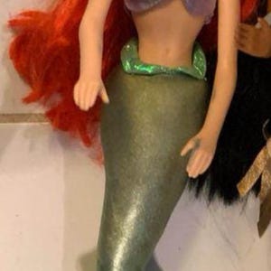 Pode incluir: Boneca de sereia com cabelo vermelho brilhante, olhos azuis e um sorriso. Ela usa um top roxo e uma cauda verde. Esta boneca &eacute; um brinquedo popular.