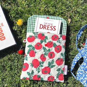 Puede incluir: Una funda acolchada para libros con un estampado de tomates, que contiene un libro titulado "The New Dress". La funda tiene un forro de cuadros verdes y un cierre de botón rojo. Un pequeño adorno con un tomate rojo cuelga del lateral.