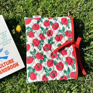 May include: Quilted fabric book cover with a white background and a pattern of red tomatoes and green leaves. The cover has a red trim and a red ribbon tie. A book with the title "Culture Handbook" is partially visible.