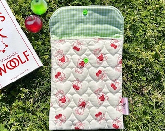 May include: A quilted cream-colored case with a heart pattern featuring red teddy bears and cherries. The top flap is green gingham, secured with a green snap. A book and two decorative glass fruits are nearby.