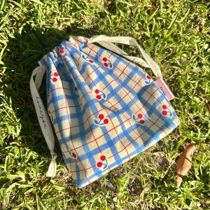 以下が含まれることがあります： 赤いチェリー柄の小さな青と白のギンガムチェックの巾着袋。袋には白い紐が付いており、紐には「love you」の文字が書かれています。