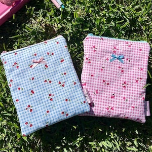 Könnte beinhalten: Zwei rechteckige Stoffbeutel mit Vichy-Muster und kleinen roten Erdbeermotiven. Einer ist hellblau, der andere rosa. Jeder hat eine kleine Schleife und einen Reißverschluss. Die Beutel liegen auf grünem Gras.