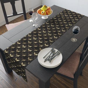 Puede incluir: Una mesa de comedor negra está decorada con un camino de mesa decorativo con un patrón de hojas doradas. Un cuenco blanco con manzanas está en el centro. Copas de vino, platos y cubiertos completan el conjunto.