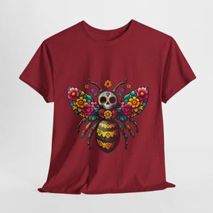 May include: Red t-shirt with a colorful sugar skull bee design. The bee has yellow and black stripes and is surrounded by pink, yellow, and orange flowers.