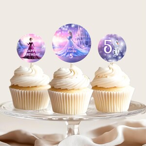 Pu&ograve; includere: Tre cupcake con glassa bianca sono esposti su un'alzatina di vetro. Ogni cupcake ha un topper circolare con personaggi e testo del film "Frozen". I topper recitano "Happy Birthday", "Amelia is turning 5" e "5" con Olaf.