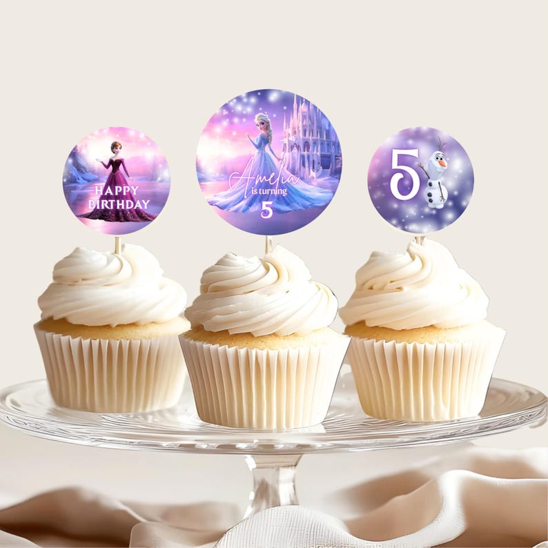 Pu&ograve; includere: Tre cupcake con glassa bianca sono esposti su un'alzatina di vetro. Ogni cupcake ha un topper circolare con personaggi e testo del film "Frozen". I topper recitano "Happy Birthday", "Amelia is turning 5" e "5" con Olaf.
