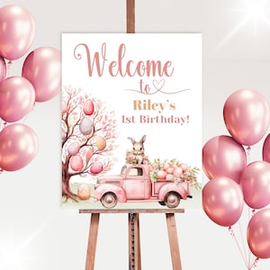 Puede incluir: Bienvenido al 1er cumpleaños de Riley! Un cartel de bienvenida rosa y dorado con una ilustración de acuarela de un camión rosa que lleva un conejito y huevos de Pascua.