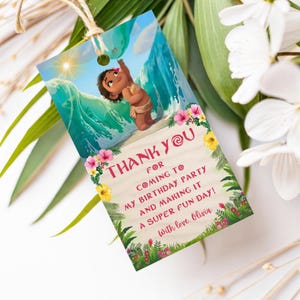 A rectangular baby Moana themed thank you tag hanging from a rope, showing a smiling baby girl standing on a beach with ocean waves behind her. The tag has tropical flowers in pink and yellow at the bottom and reads “Thank you for coming to my birthday party and making it a super fun day.”