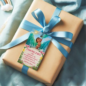 A beige gift box wrapped with a soft blue ribbon, decorated with a hanging baby Moana themed thank you tag. The tag shows a smiling baby girl standing on a beach with ocean waves, sunlight, and tropical flowers. The message on the tag thanks guests for coming to the birthday party.