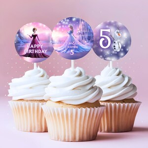 Pu&ograve; includere: Tre cupcake con glassa bianca, ognuno sormontato da un'immagine circolare su uno stecchino. Le immagini presentano personaggi e testo del film "Frozen", tra cui Elsa, Anna, Olaf e il numero 5. Il testo include "Buon Compleanno" e un nome.