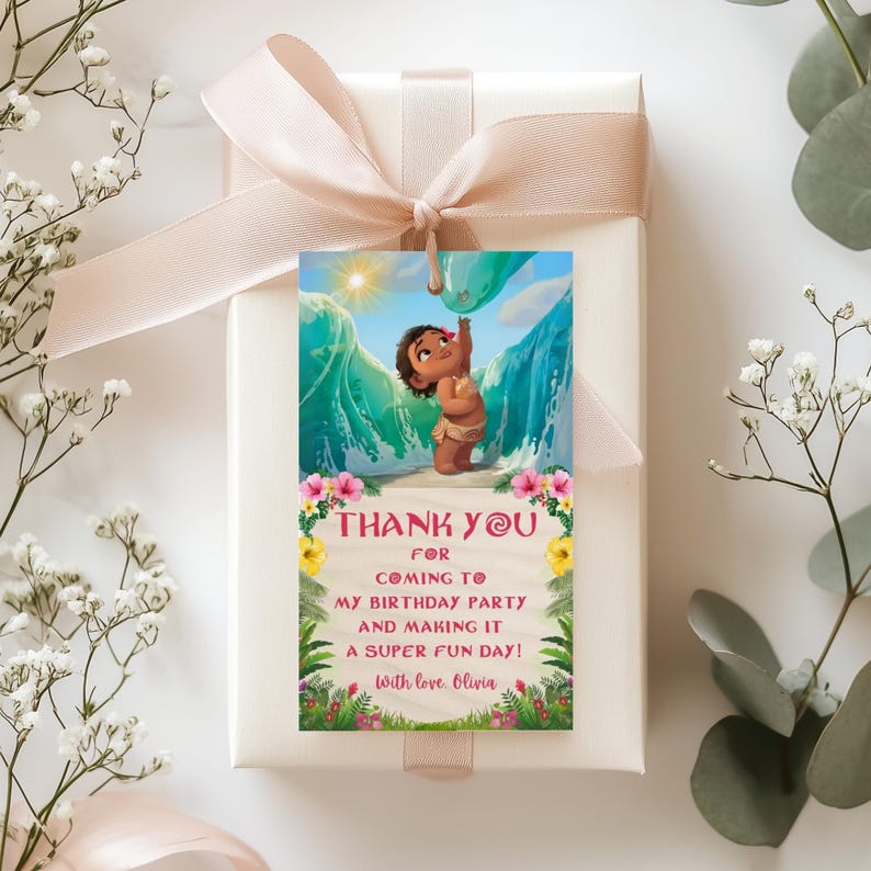 A close-up of a gift box wrapped in beige paper with a soft ribbon, decorated with a hanging baby Moana themed thank you tag. The tag includes floral decorations and a warm thank you message.