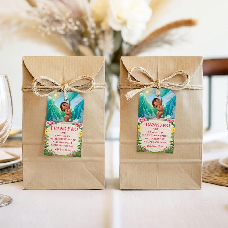 Two kraft paper gift bags placed side by side on a table, each tied with string and decorated with a baby Moana favor tag. The tags show a tropical ocean background and a cheerful thank you message.